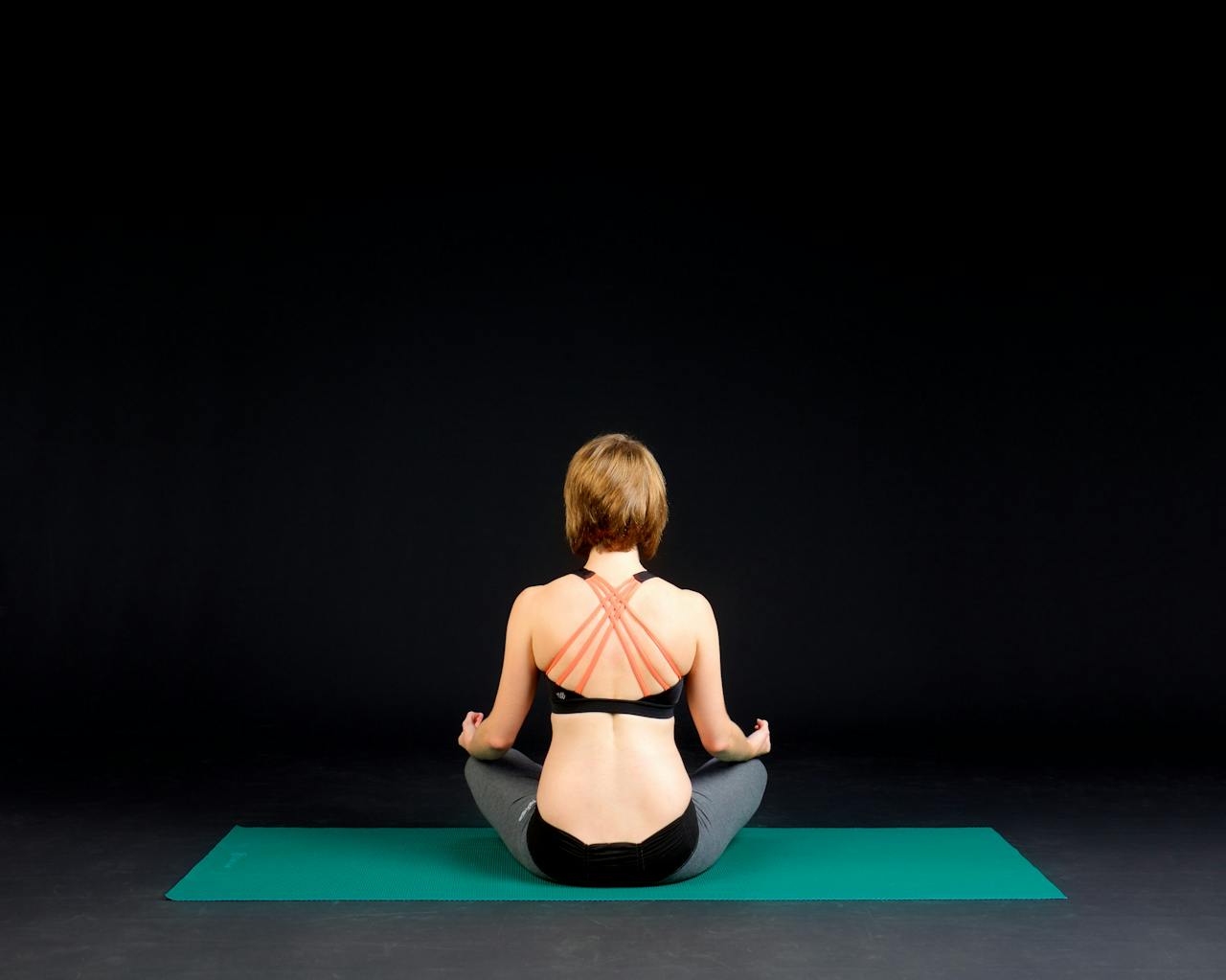 Back view of a woman meditating on a mat in a dark room, promoting relaxation and mindfulness.